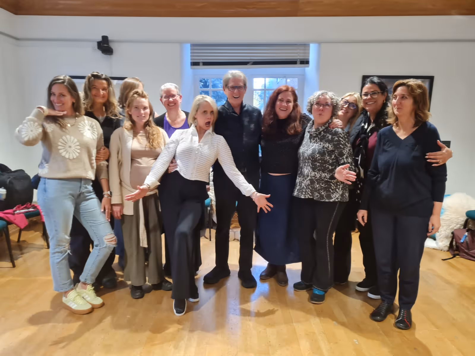 Group of twelve women standing indoors, some smiling and posing playfully in front of a window with blinds.