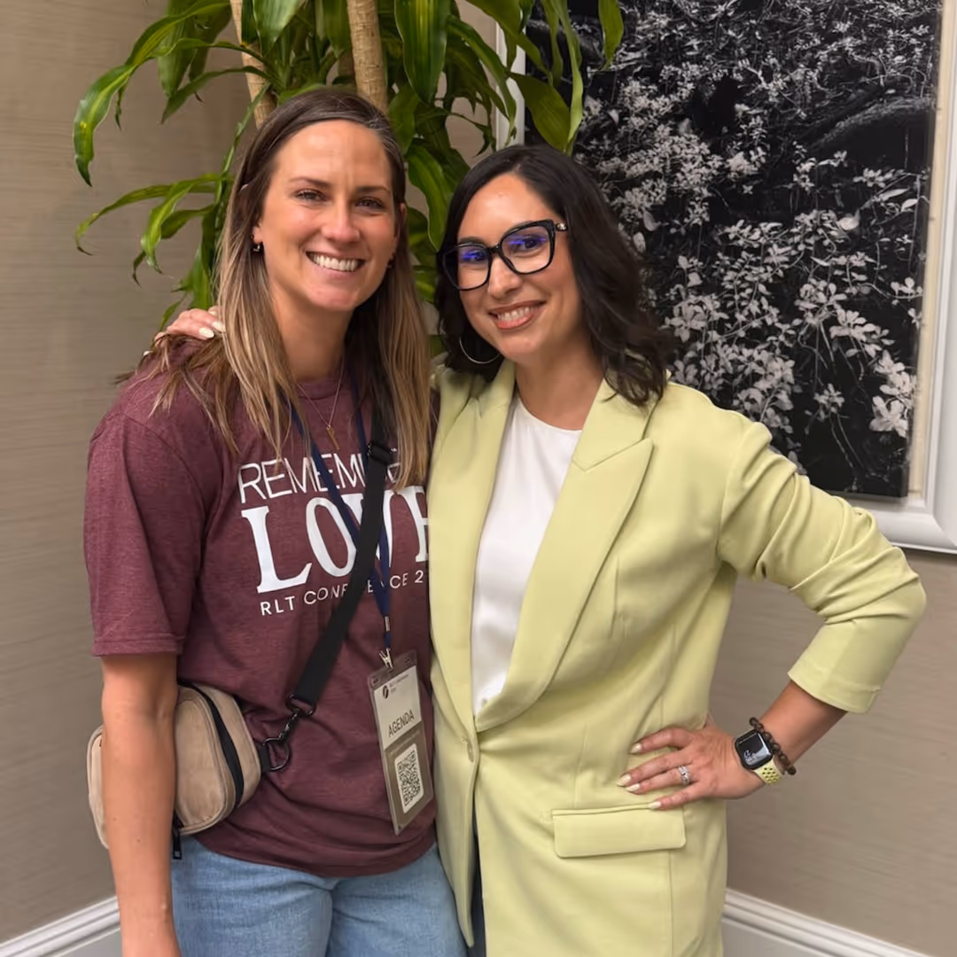 Two women smiling and posing together indoors, one wearing glasses and a pale yellow blazer, the other in a maroon T-shirt with a conference badge.