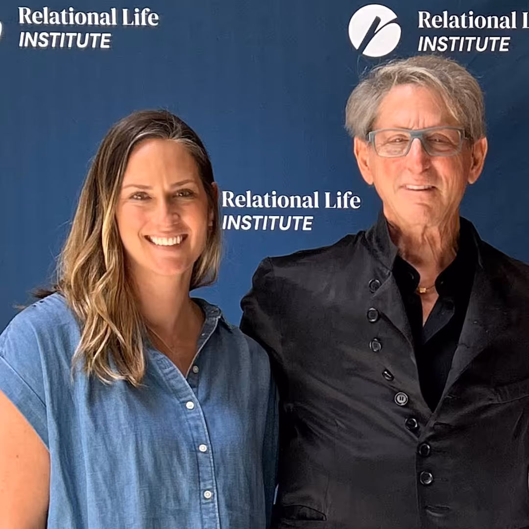 Smiling man in glasses wearing a black jacket and a woman in a blue button-up shirt standing in front of a Relational Life Institute backdrop.
