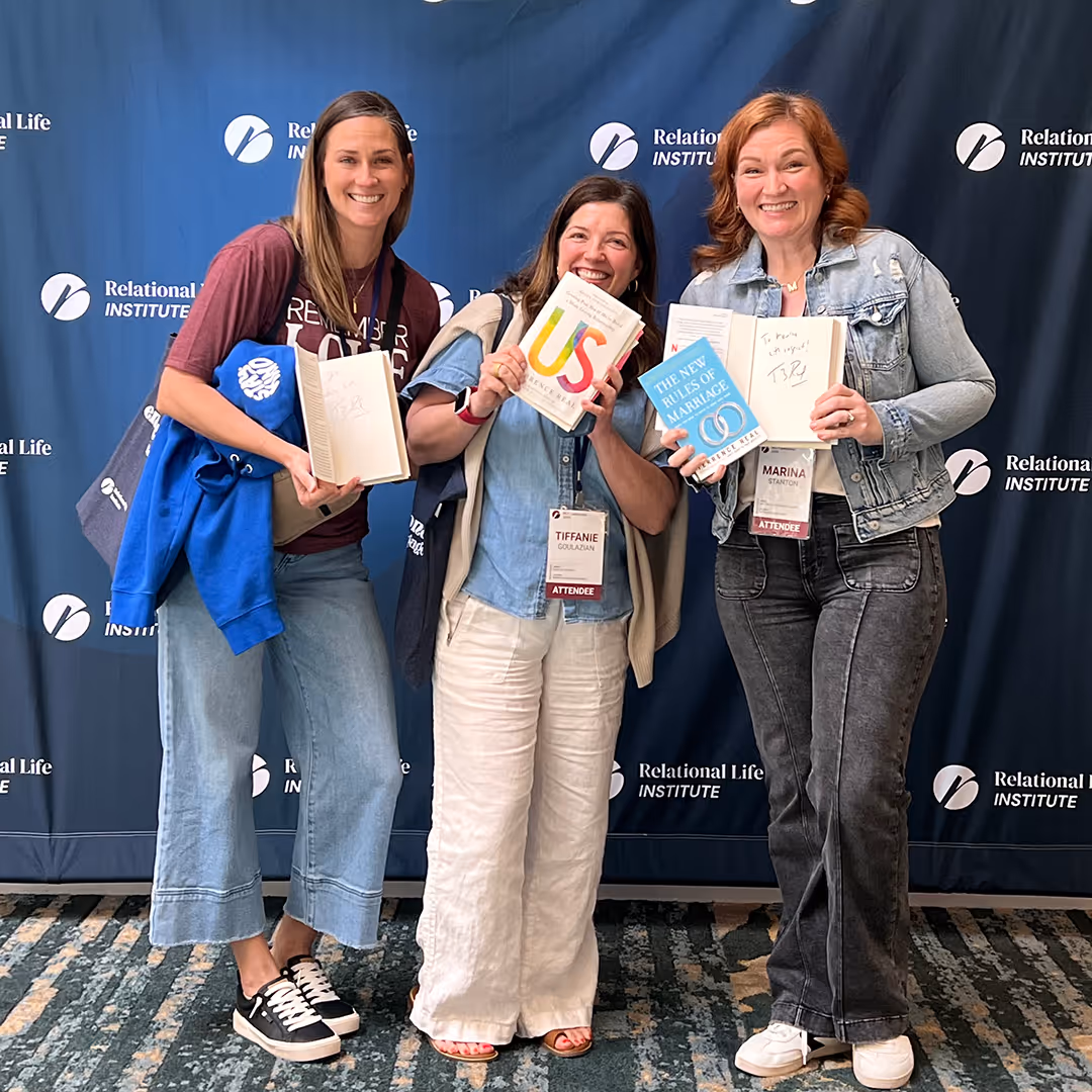 Three smiling women holding signed books in front of a Relational Life Institute backdrop at an event.