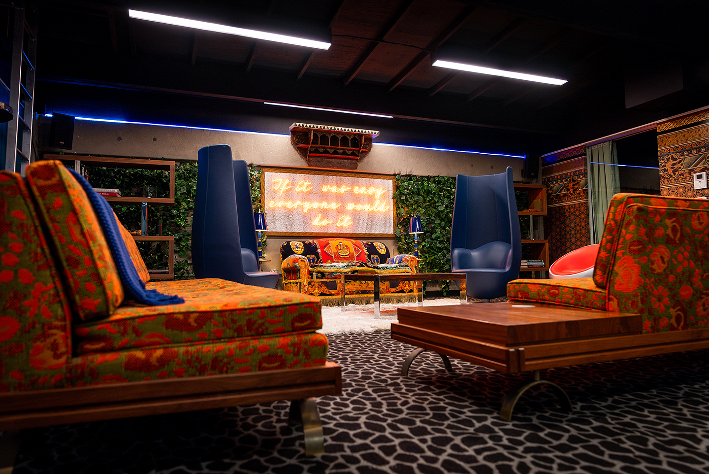 Lounge area with vibrant patterned sofas, two tall blue chairs, a wooden coffee table, green wall, and neon sign saying 'If it was easy everyone would do it.'