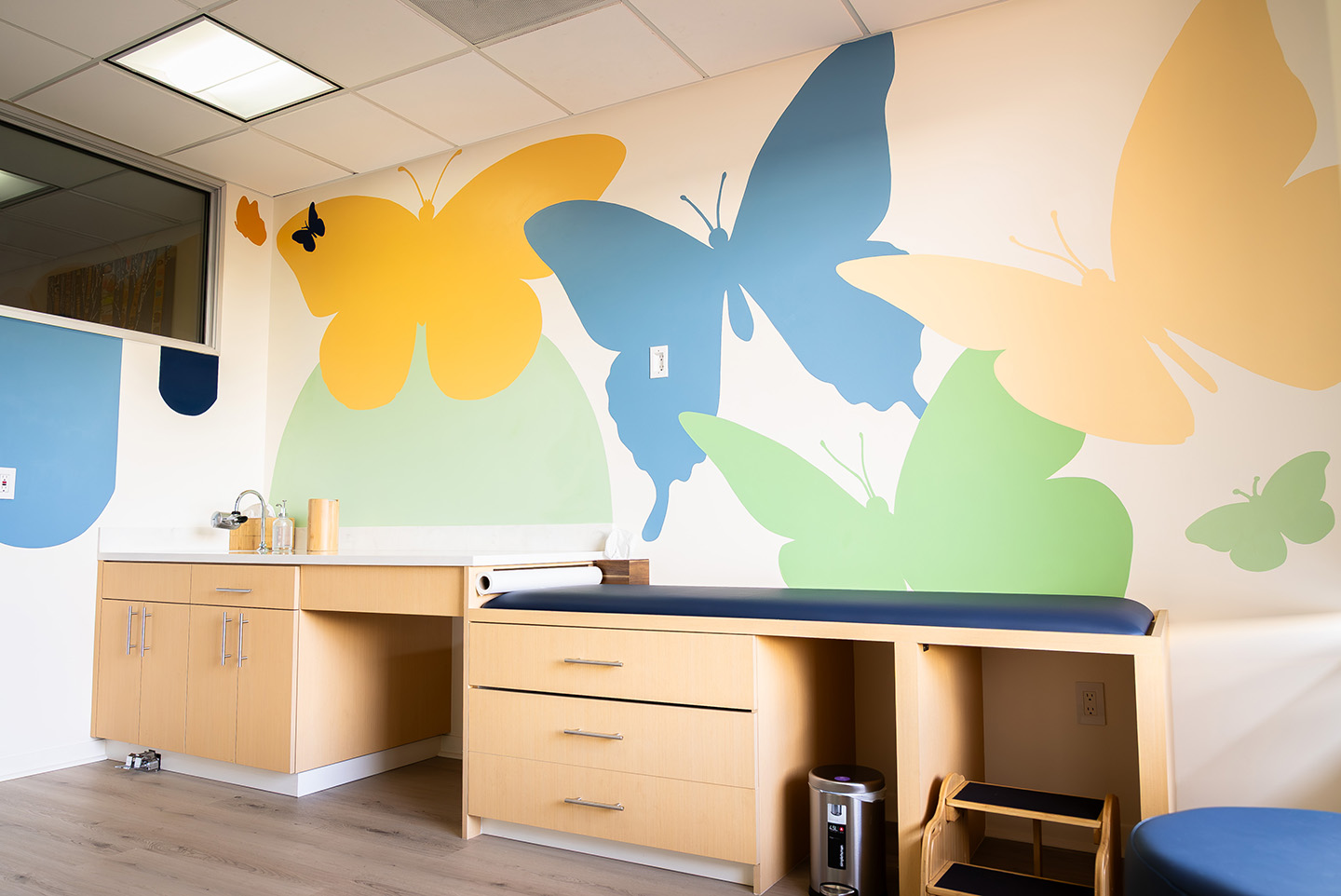 Bright medical exam room with butterfly mural on the wall, light wood cabinets, and a blue padded exam table.