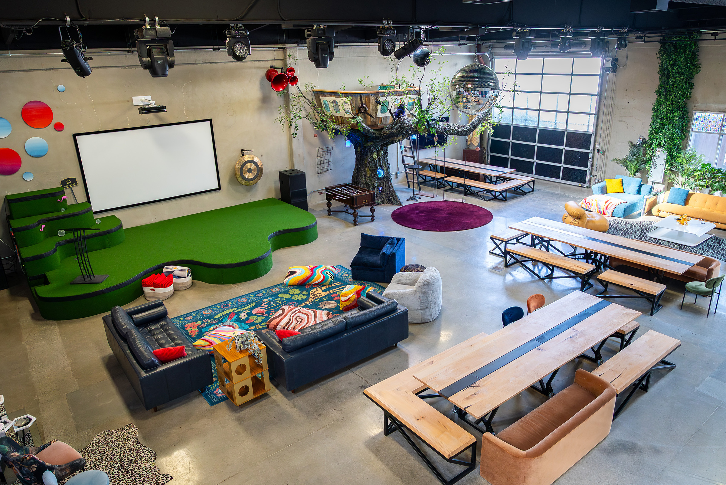Spacious modern event space at FutureHouse, a San Francisco AI company, featuring a custom stage, large screen, colorful seating areas, custom wooden tables with benches, and a fabricated treehouse with a disco balls, designed and built by FrozenFeathers.
