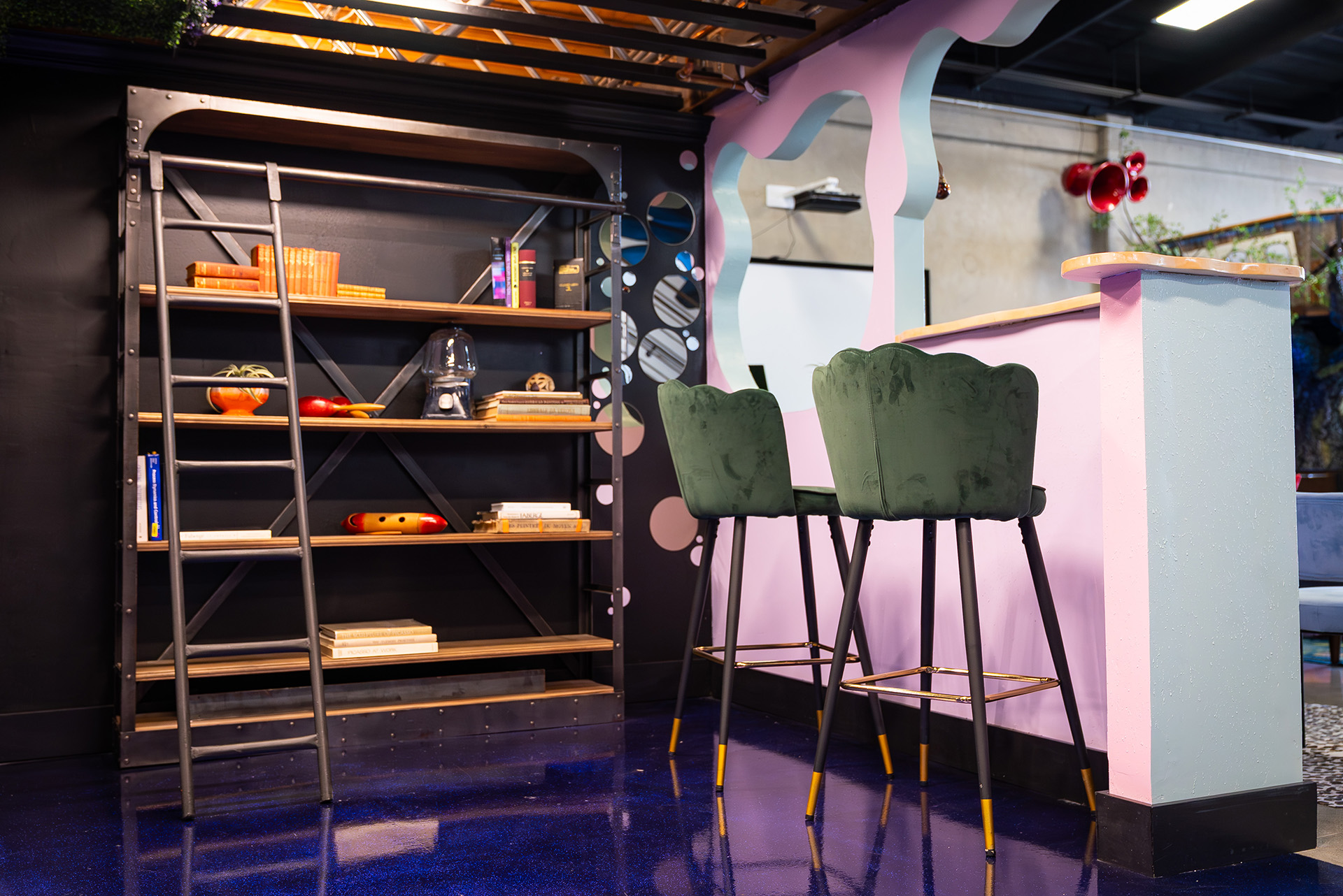 “Modern game room at FutureHouse in San Francisco featuring a black metal bookshelf, colorful books, green velvet bar stools, and a pink and light blue bar counter, designed and built by FrozenFeathers.