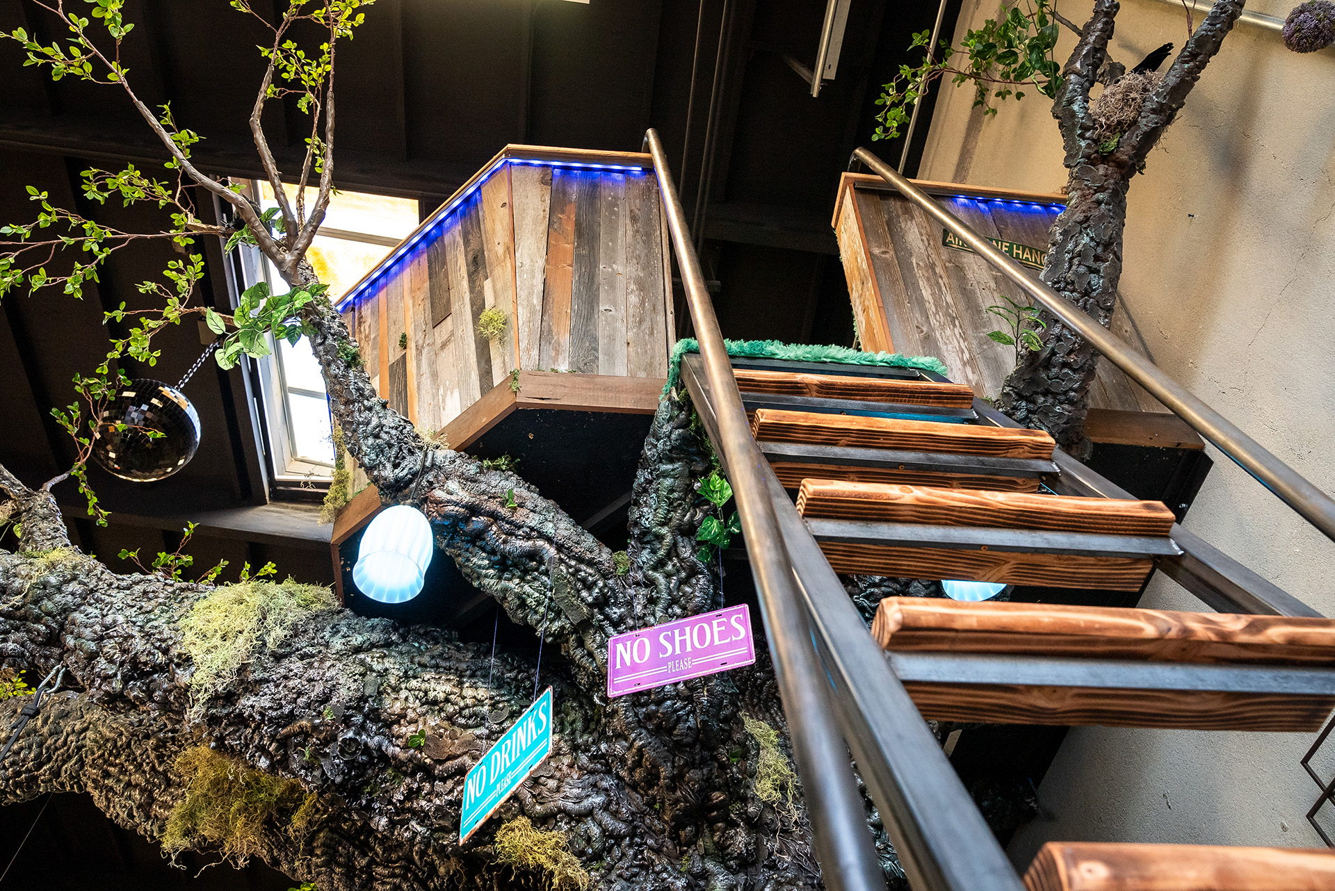 Custom-fabricated treehouse staircase with wood steps and metal railings leading to a handcrafted loft structure, framed by sculptural branches and “No Shoes” and “No Drinks” signs, built for FutureHouse.