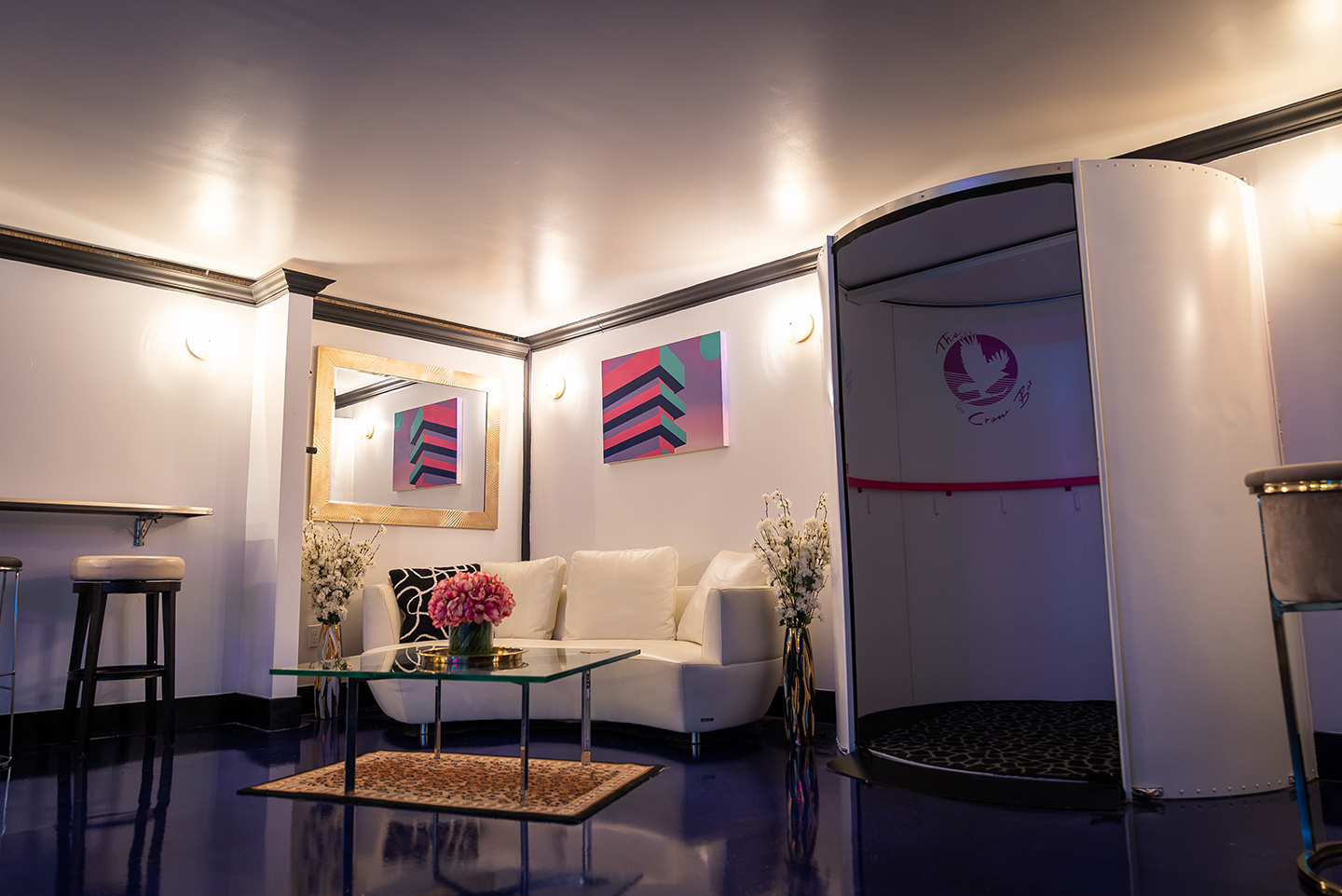Speakeasy lounge interior with a white curved sofa, glass coffee table with pink flowers, tall floral vases, bar stools, wall art, and a circular white photo booth.