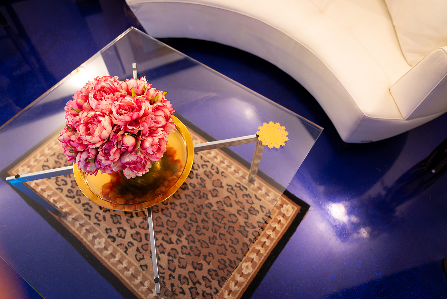Pink peony bouquet in a golden vase on a glass coffee table over a leopard print rug next to a cream curved sofa on shiny blue floor.