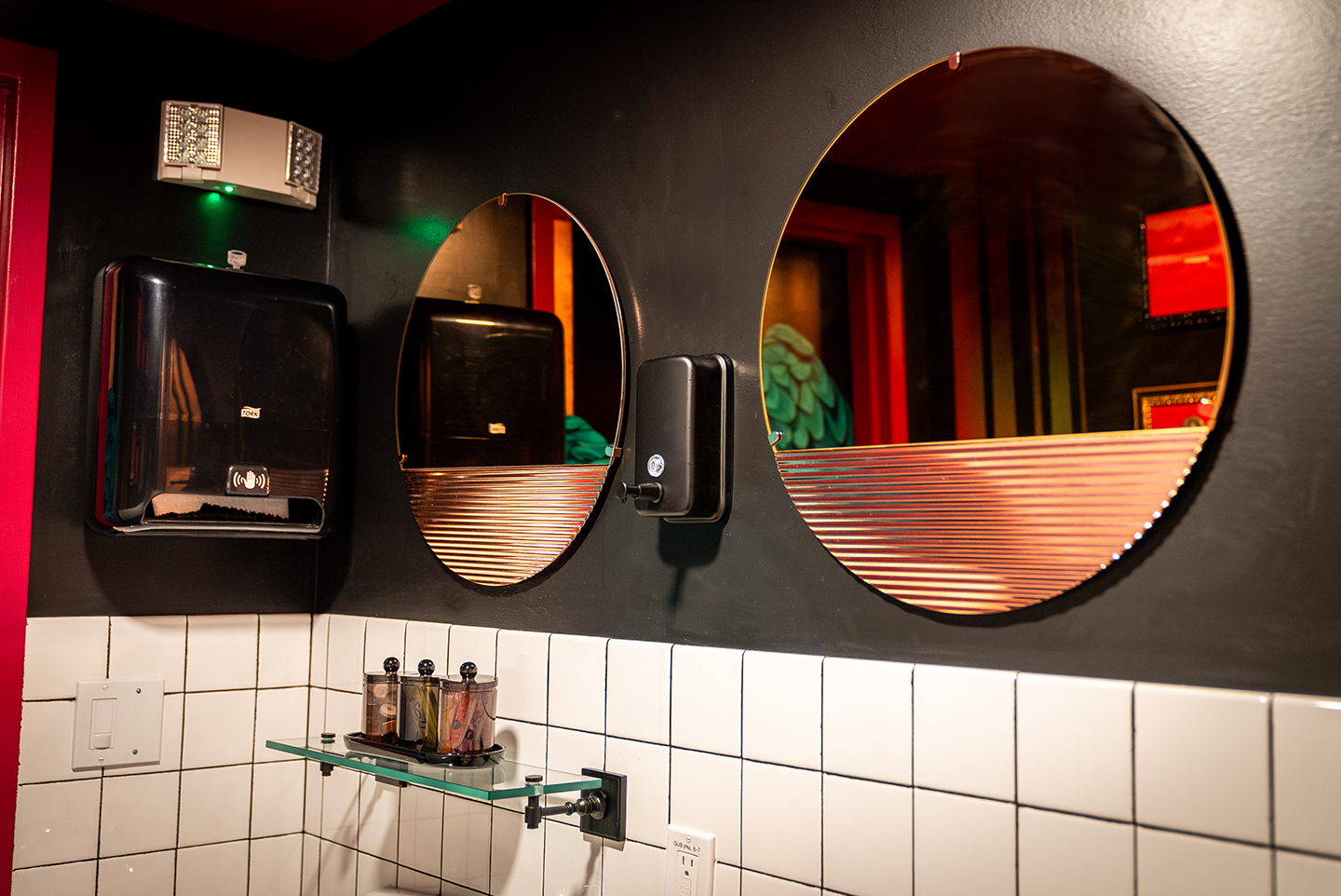 Bathroom interior with black walls, two round mirrors with horizontal striped design, a black soap dispenser, a black paper towel dispenser, and a glass shelf holding three small containers.