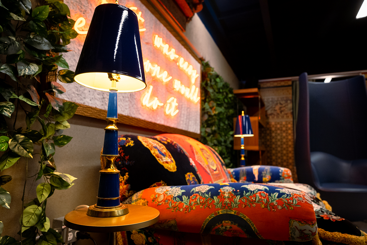 Blue and gold table lamps beside a colorful patterned sofa with floral and ornate designs, against a wall with green vines and a neon sign in a cozy indoor setting.