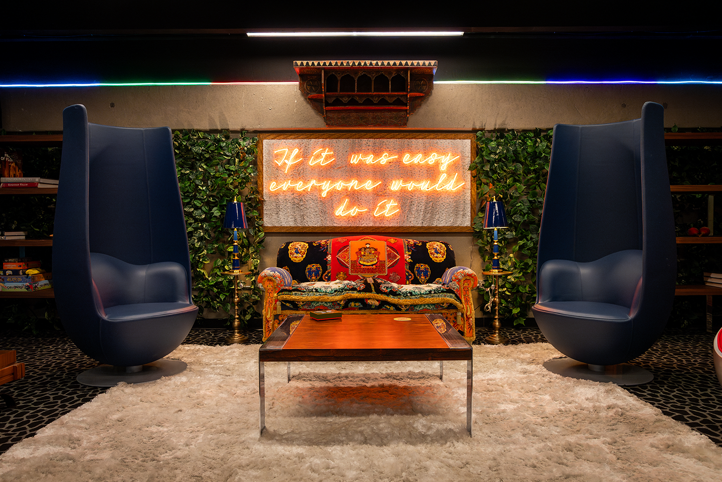 Commercial lounge interior with tall blue armchairs, a colorful patterned sofa, wooden coffee table, fluffy rug, and a neon sign reading “If it was easy everyone would do it.”