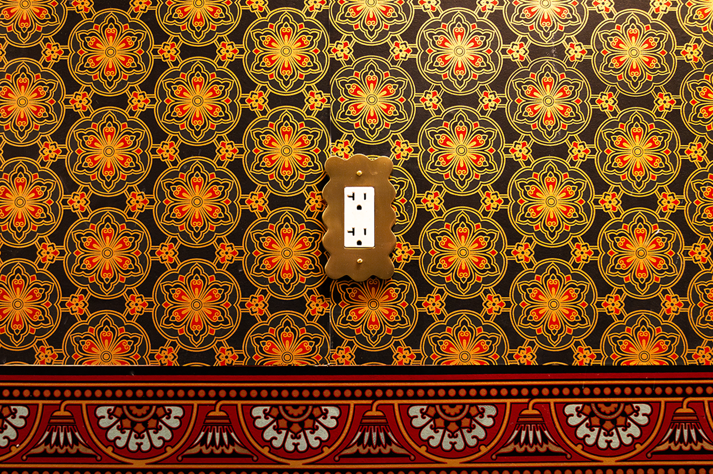 Electric outlet with decorative brass cover mounted on a wall with intricate orange, yellow, and black floral patterned wallpaper and red ornamental border at the bottom.