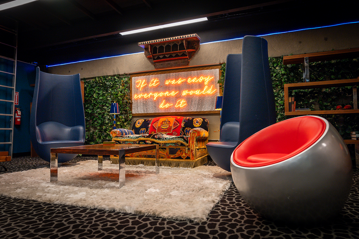 Modern Versace-inspired lounge featuring a patterned sofa, tall blue chairs, a round silver chair with a red cushion, and a neon sign reading “If it was easy everyone would do it.