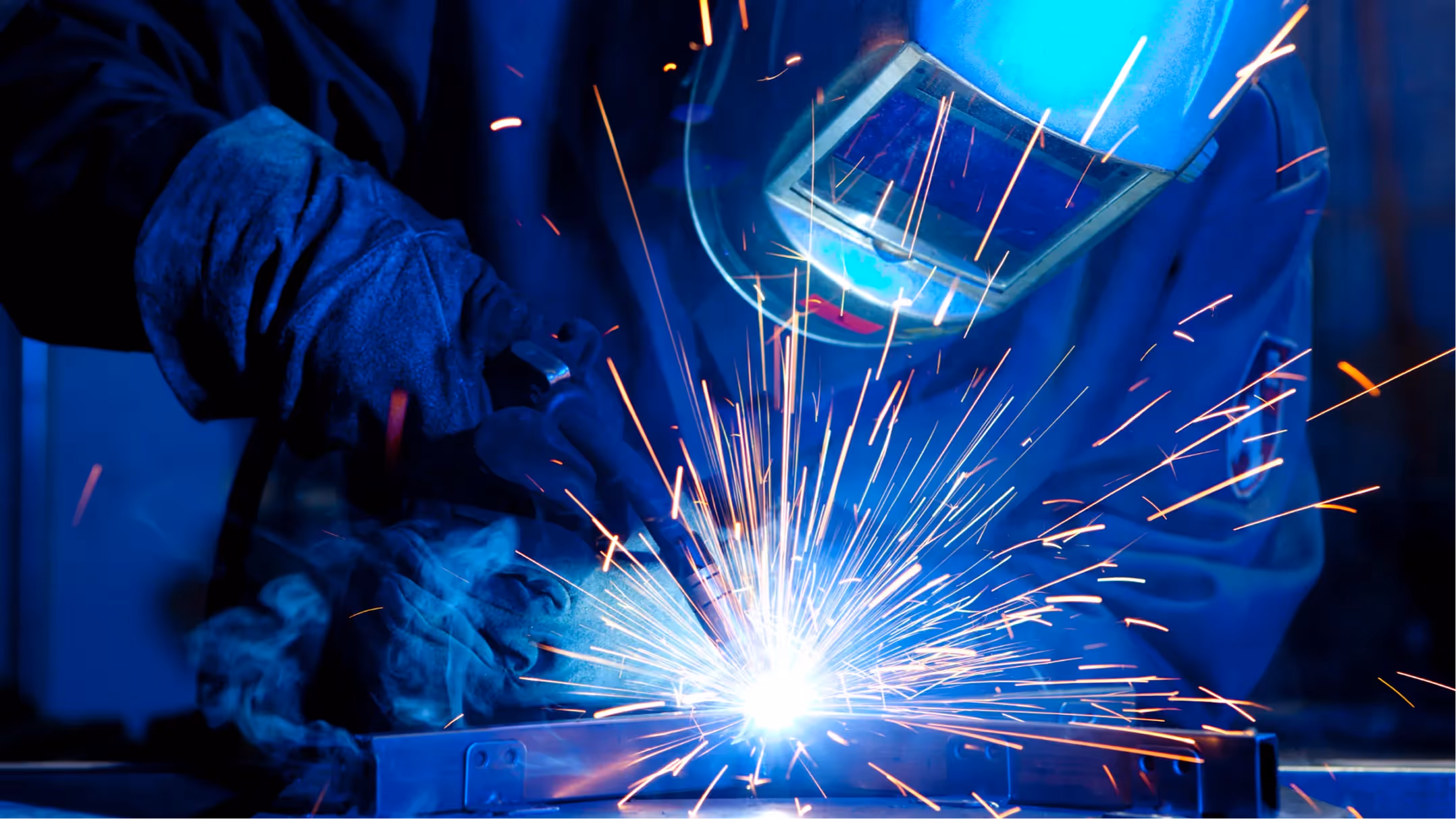Welder wearing protective gloves and helmet working with bright sparks flying from welding metal.