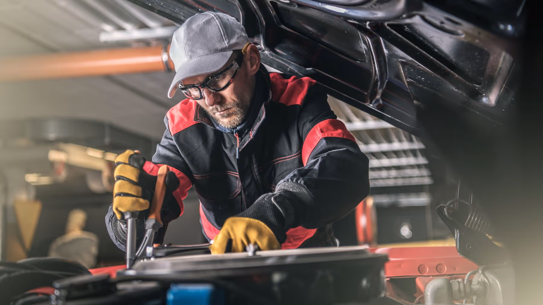 Mechanic wearing safety glasses and gloves working under the open hood of a car.