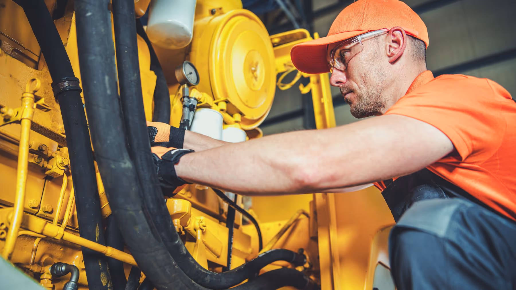 Male technician in orange cap and shirt inspecting large yellow industrial machinery with black hoses.