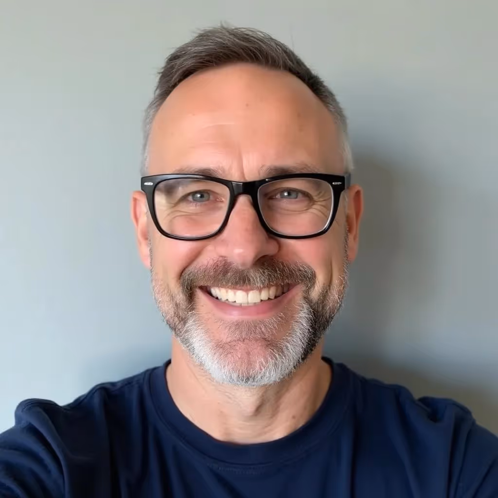 Middle-aged man with short gray hair, black glasses, and beard smiling against a plain light gray background.