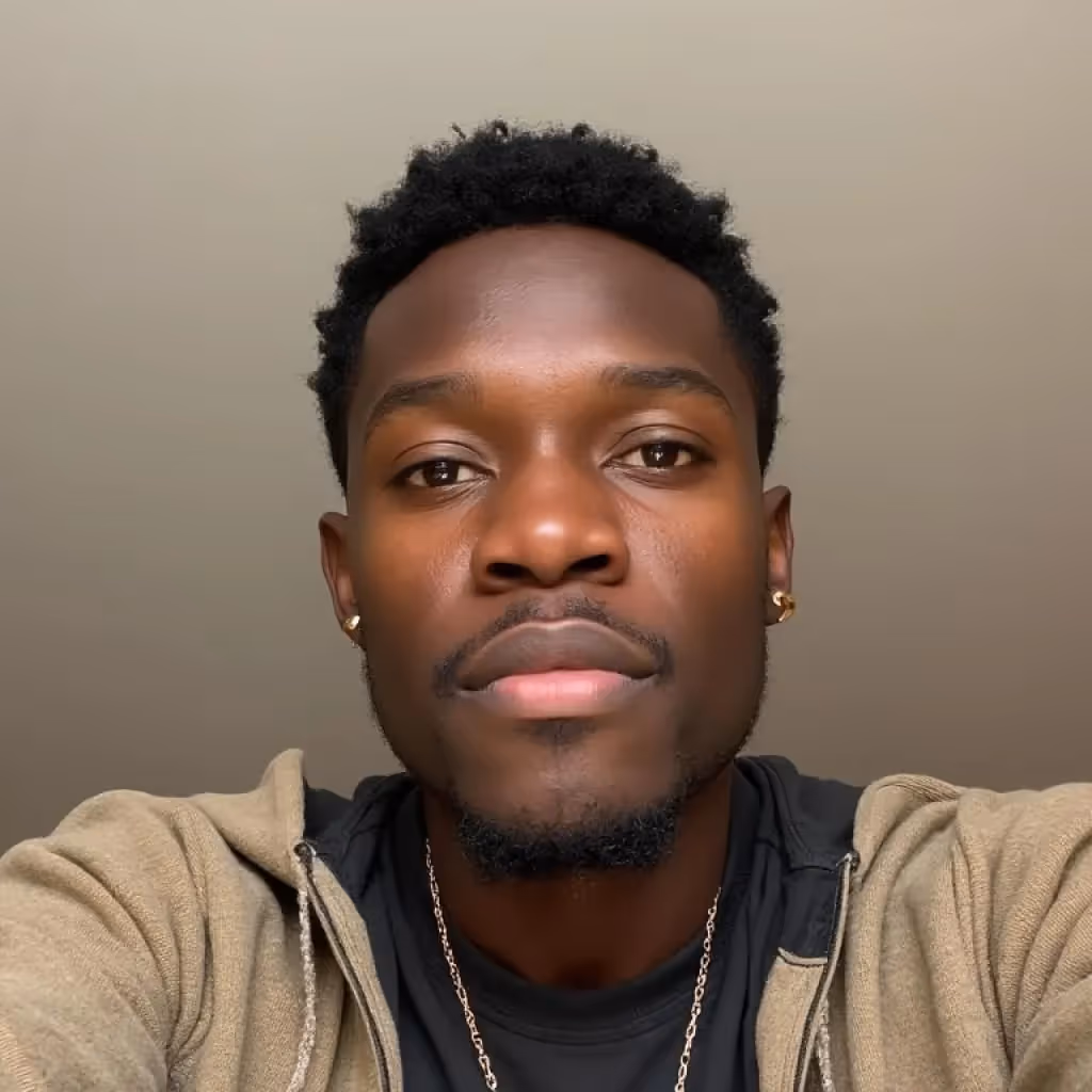 Close-up of a man with short curly hair, wearing gold earrings, a gold chain, a black shirt, and a beige jacket against a neutral background.