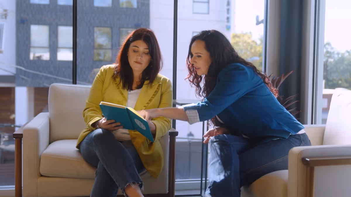 Two women sitting on separate chairs by a large window, one reading a notebook while the other points at it.