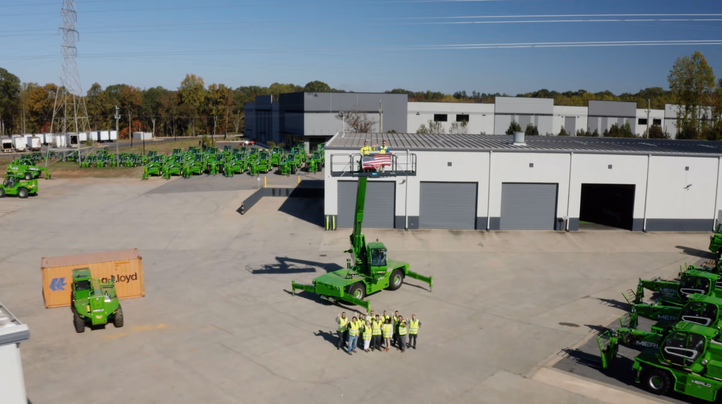 A group of workers in yellow safety vests waving in front of a green telescopic handler lift holding two people with an American flag above a warehouse and parked green machinery.