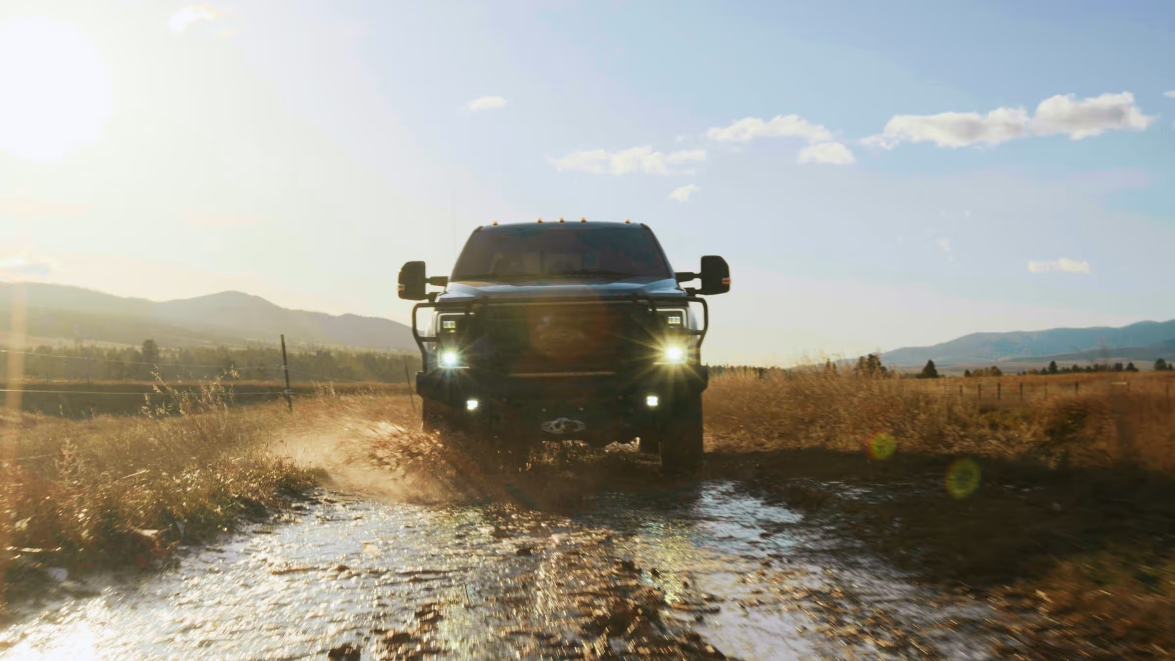 Black pickup truck driving through a muddy puddle on a rural dirt road with fields and mountains in the background at sunset.