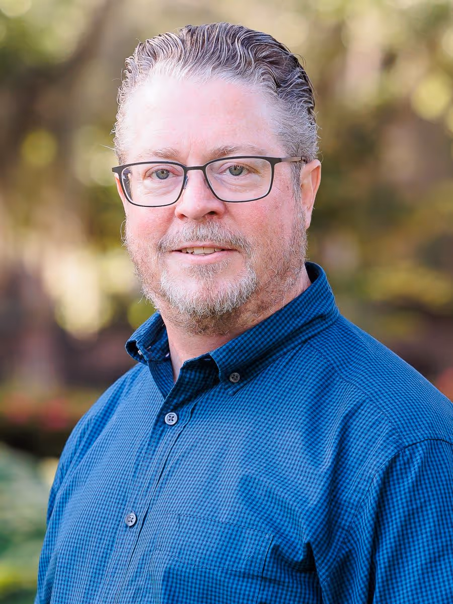 Middle-aged man with glasses, short gray hair, and beard wearing a blue checkered shirt outdoors.