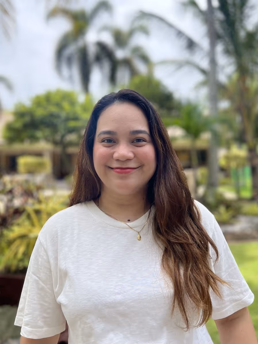 Smiling woman with long brown hair wearing a white shirt and gold necklace standing outdoors with blurred greenery and palm trees in the background.