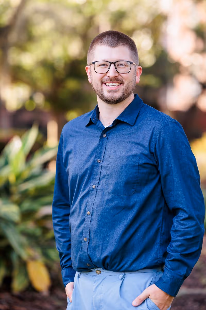 Smiling man with glasses wearing a blue shirt and light pants standing outdoors with hands in pockets.