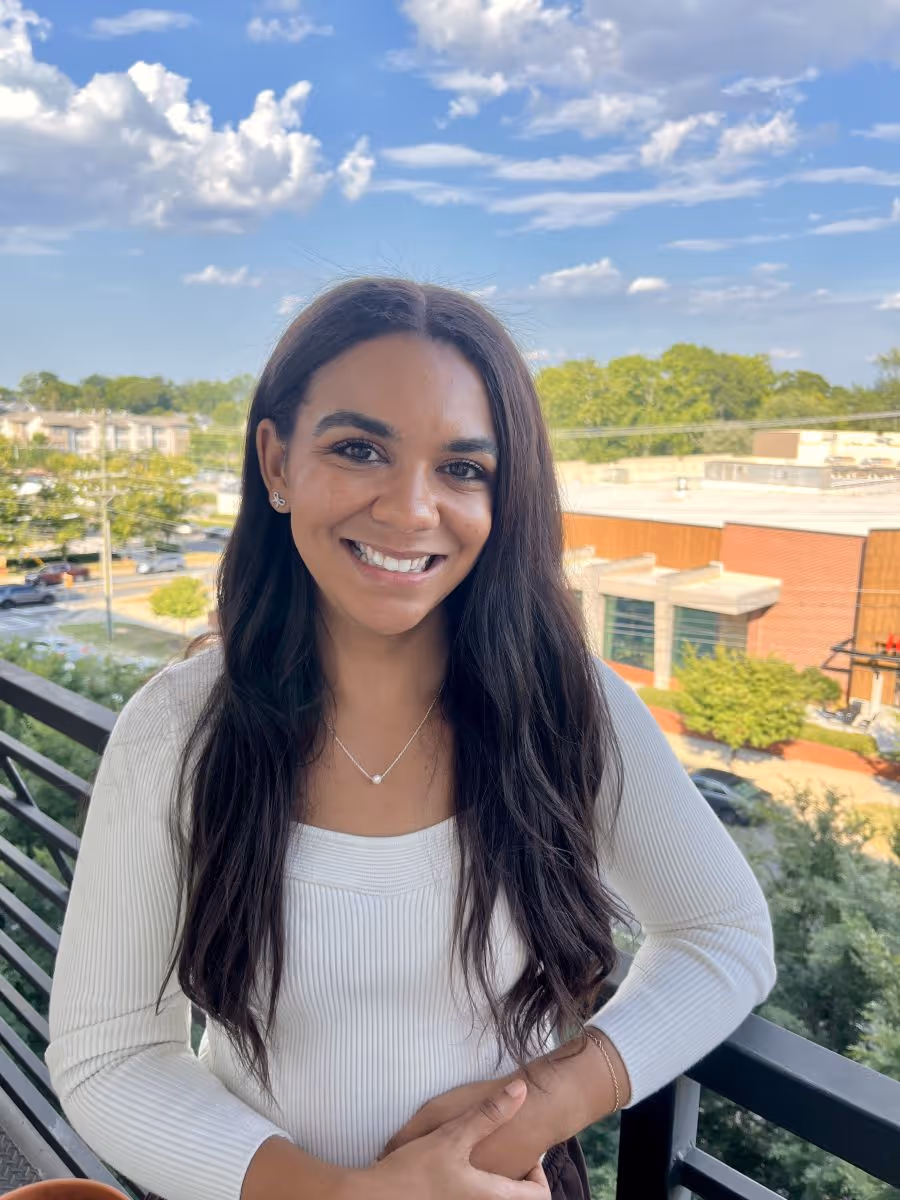 Smiling woman with long dark hair wearing a white sweater standing on a balcony with a cityscape and blue sky in the background.