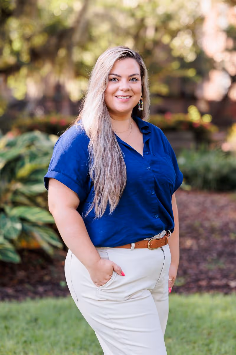 Woman with long blonde hair, wearing a blue short-sleeve shirt and beige pants, standing outdoors with hand in pocket.