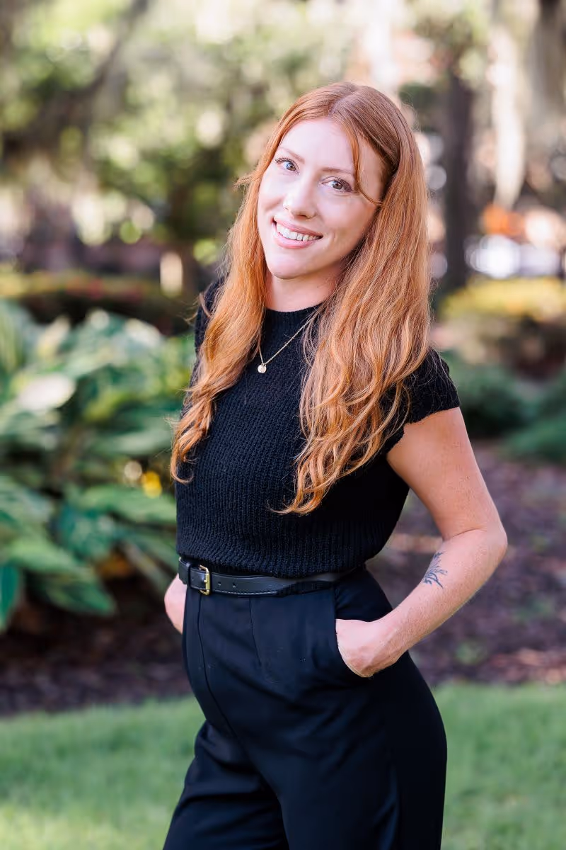 Smiling red-haired woman in a black sleeveless top and pants with hands in pockets standing outdoors.