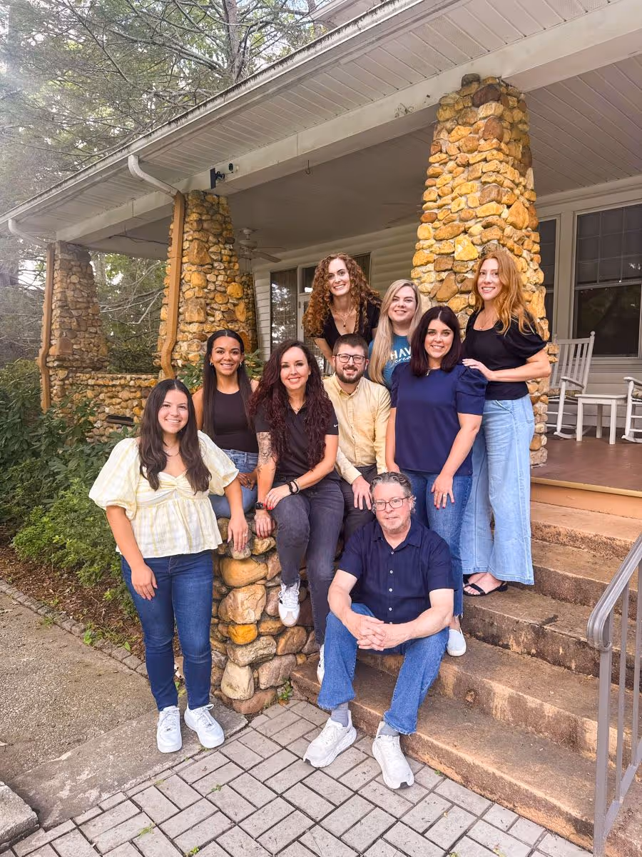Group of eight people smiling and posing on stone steps and porch of a rustic house with stone pillars.