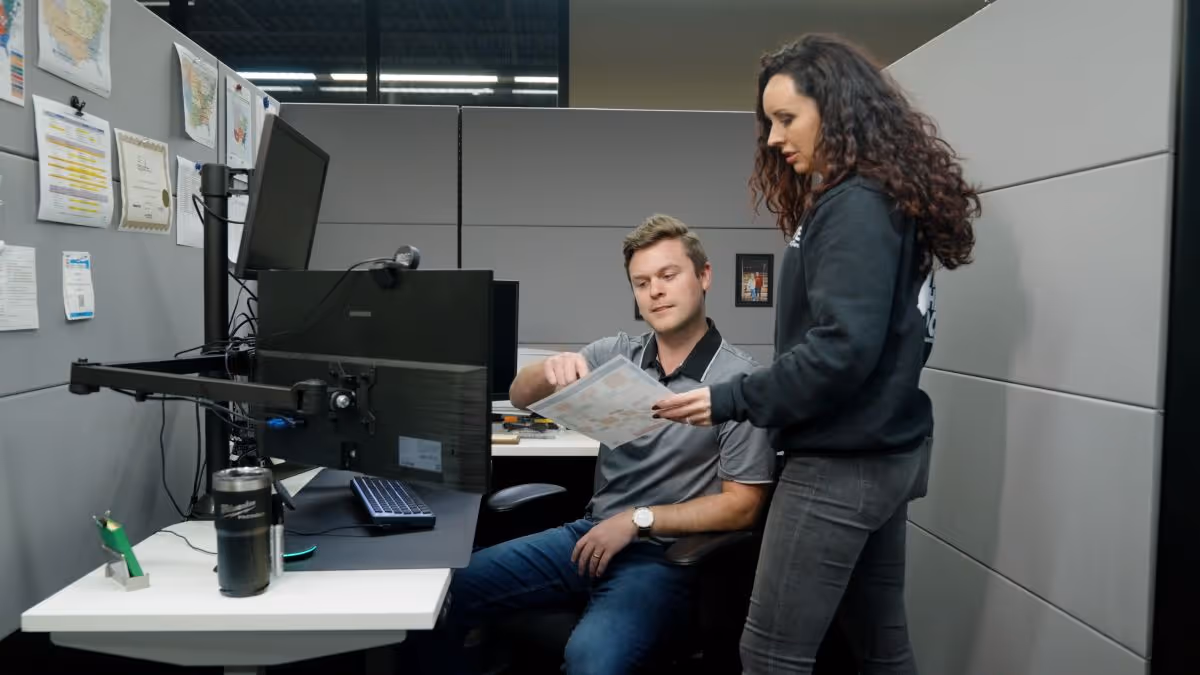Man sitting at a desk with dual monitors reviews a document handed by a standing woman in an office cubicle.