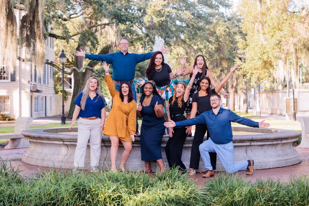 Group of nine diverse people smiling and posing enthusiastically in front of a stone fountain in a park.