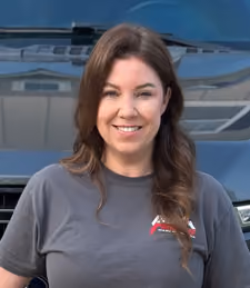 Smiling woman with long brown hair standing in front of a dark vehicle, wearing a grey T-shirt with a small logo on the left chest.