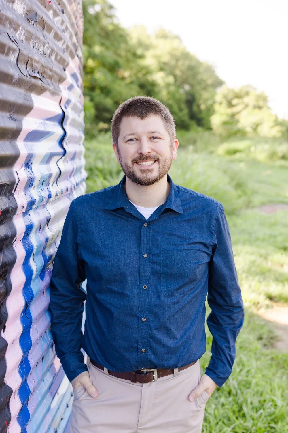 Smiling man with glasses wearing a blue shirt and light pants standing outdoors with hands in pockets.