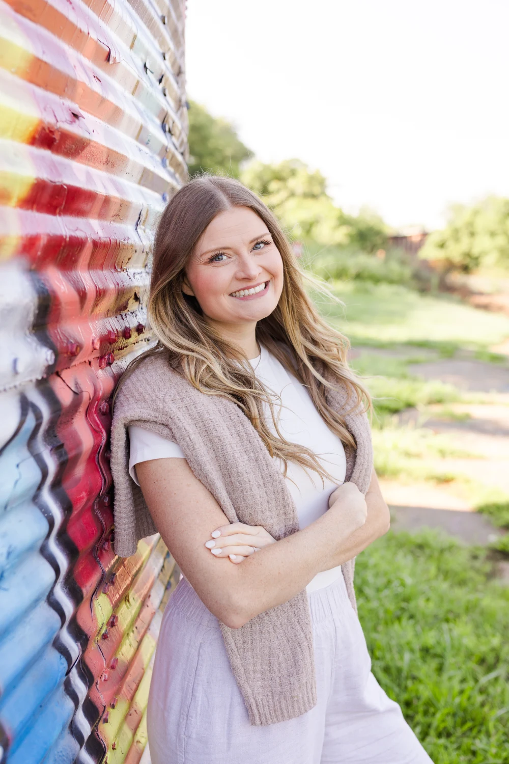 Smiling young woman with blonde hair, wearing a white t-shirt and a necklace, standing in front of a light stone wall.