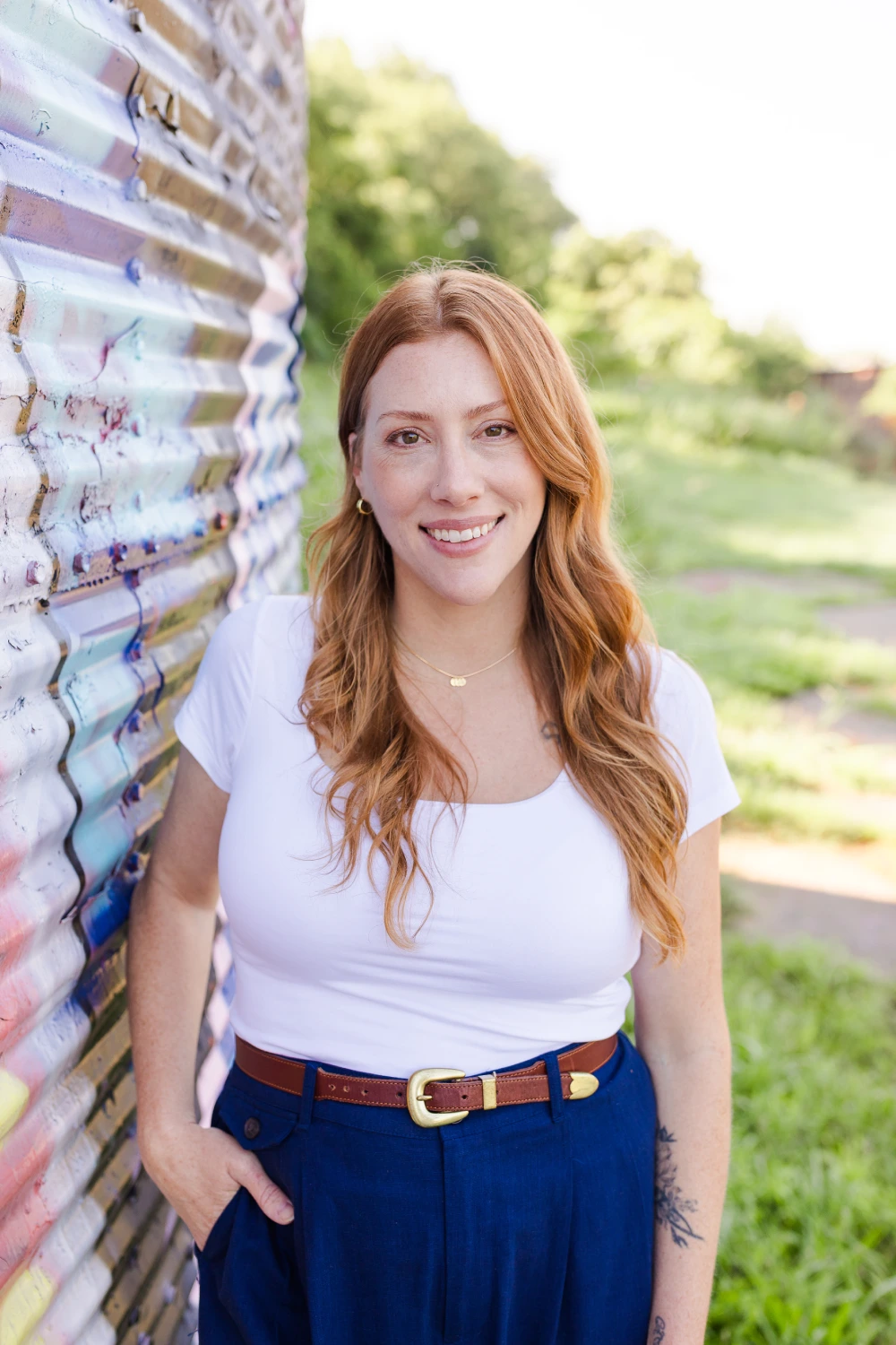 Smiling red-haired woman in a black sleeveless top and pants with hands in pockets standing outdoors.