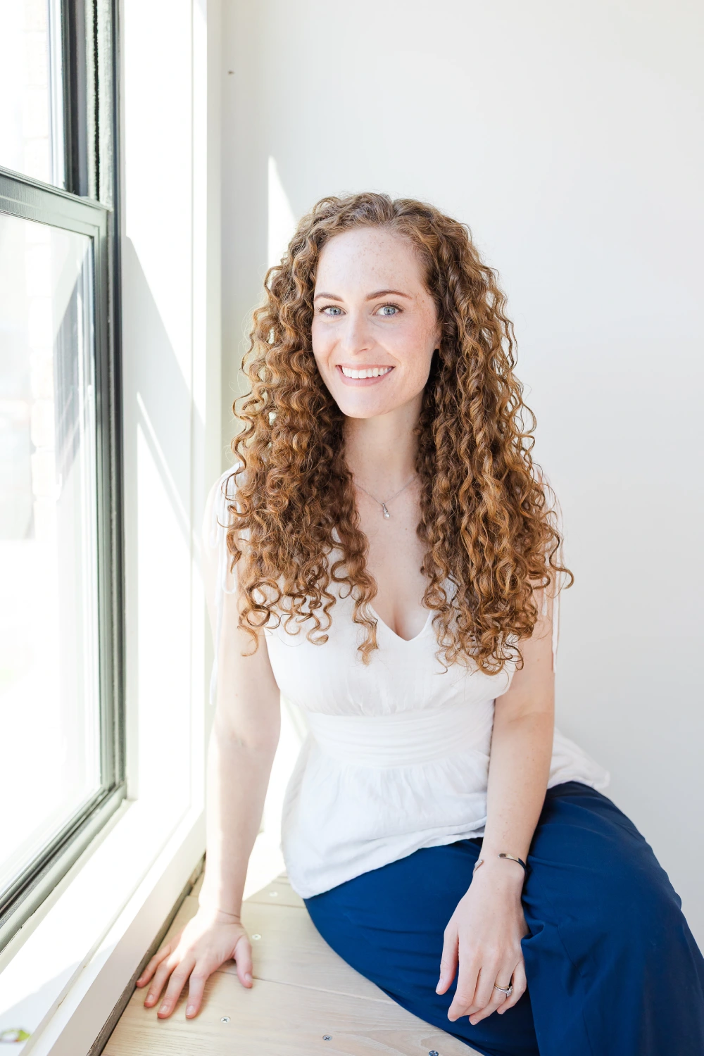 Smiling young woman with long curly red hair tied partially up, wearing a green shirt and a necklace, standing outdoors in front of a brick wall and stairs.