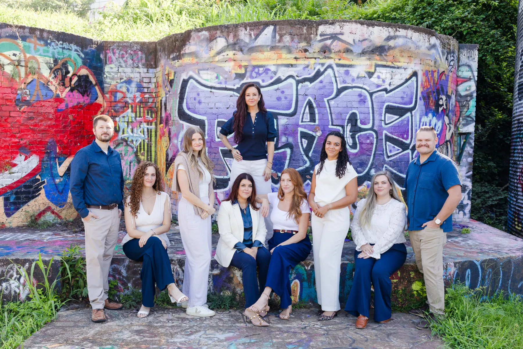 A group of nine adults standing outdoors on green grass in a park with large trees and plants in the background; they are smiling and dressed in a mix of casual and business casual attire.