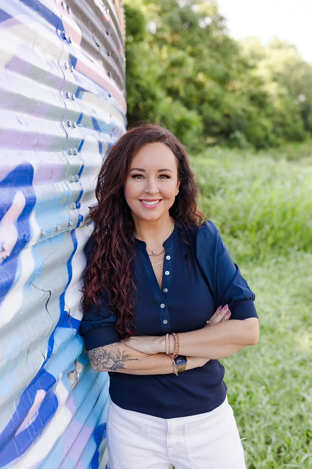 Smiling woman with long curly dark hair wearing a black short-sleeve shirt, gold chain necklace, colorful patterned skirt, and a smartwatch, standing outdoors with arms crossed in front of blurred greenery.