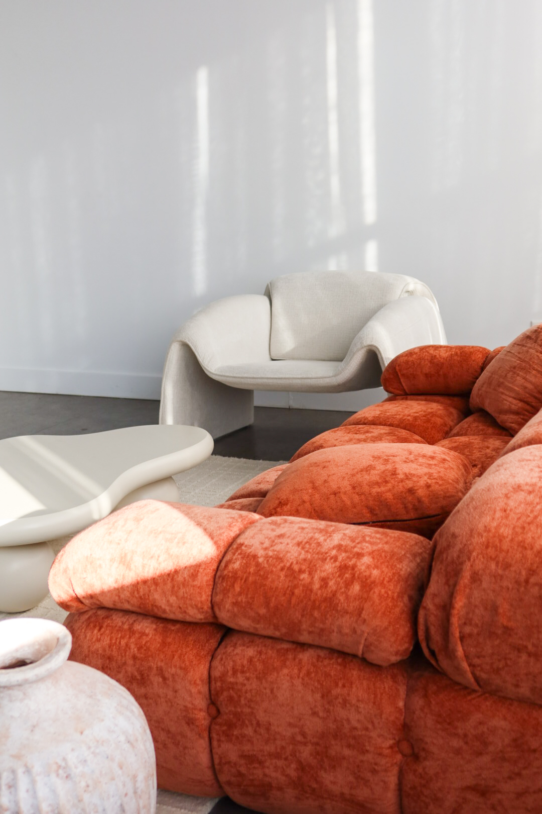 Two rustic ceramic jugs, one smaller and terracotta-colored, the other larger and off-white, standing on a tiled floor with soft natural light and shadows on a plain wall background.