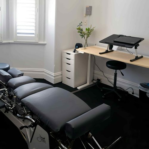 Modern chiropractic treatment room with padded adjustment table, desk, stool, and window with blinds.