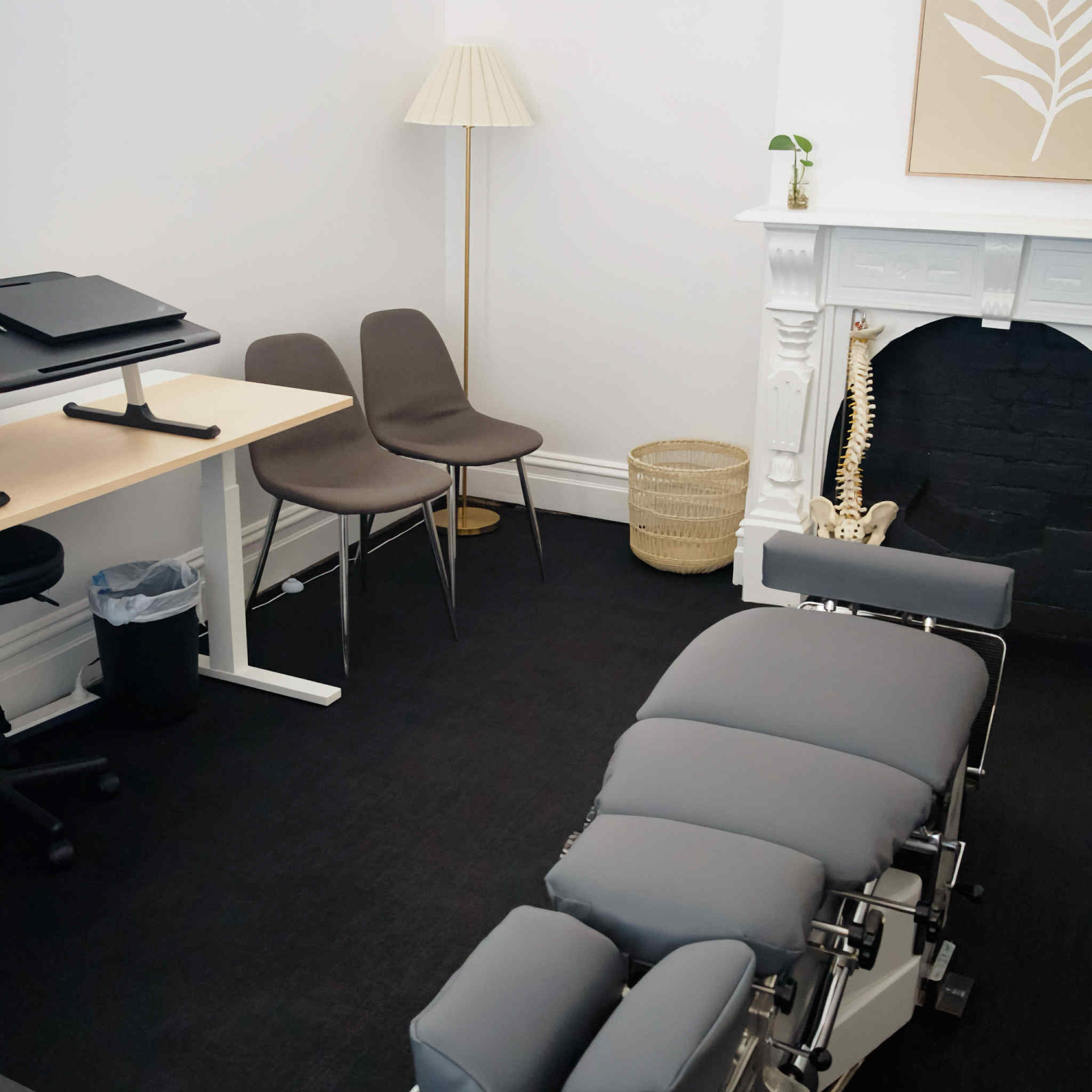 Modern chiropractic office with adjustable gray treatment table, two brown chairs, and a decorative spine model near a white fireplace.