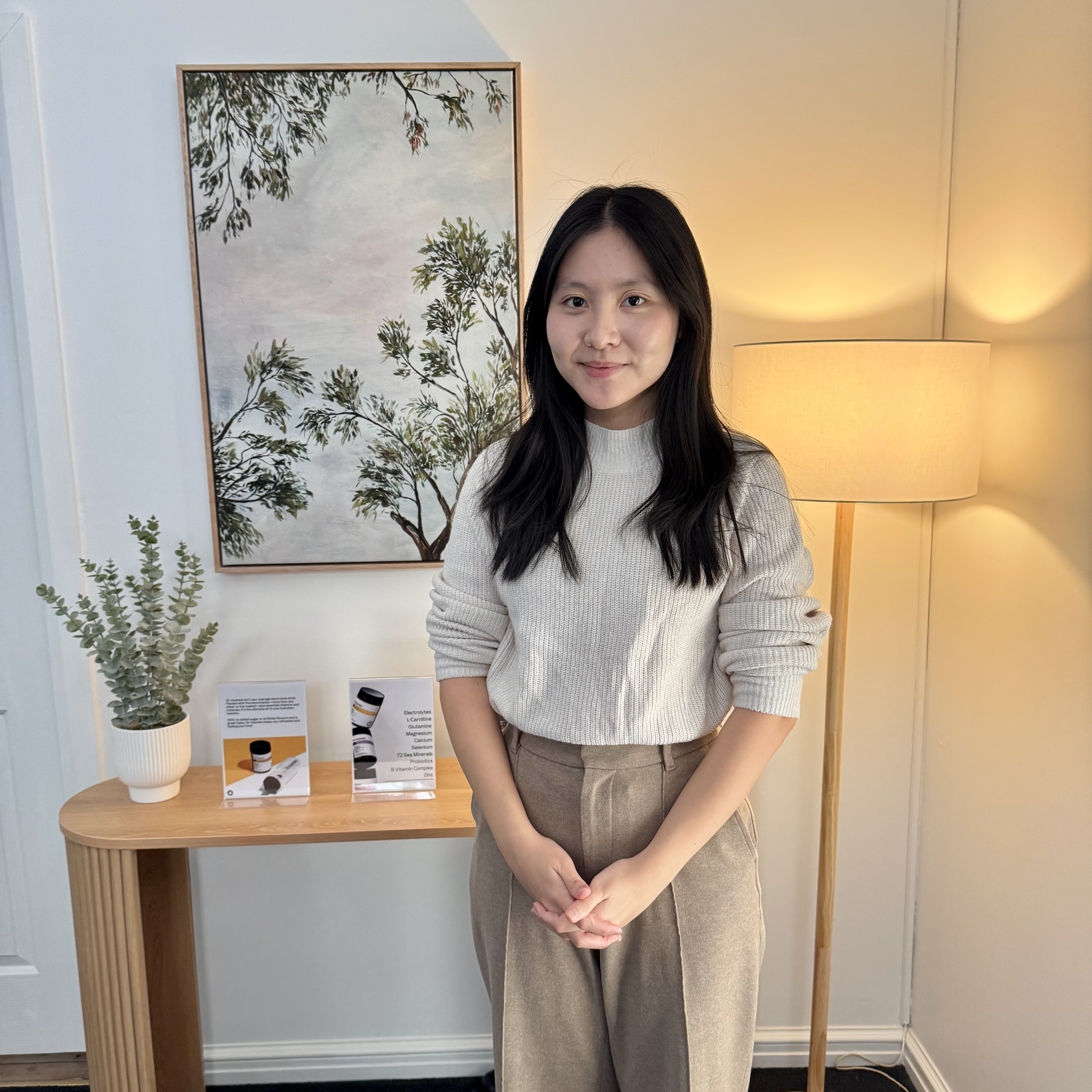 Woman with black hair in beige sweater and pants standing in a room with a wooden table and lamp.