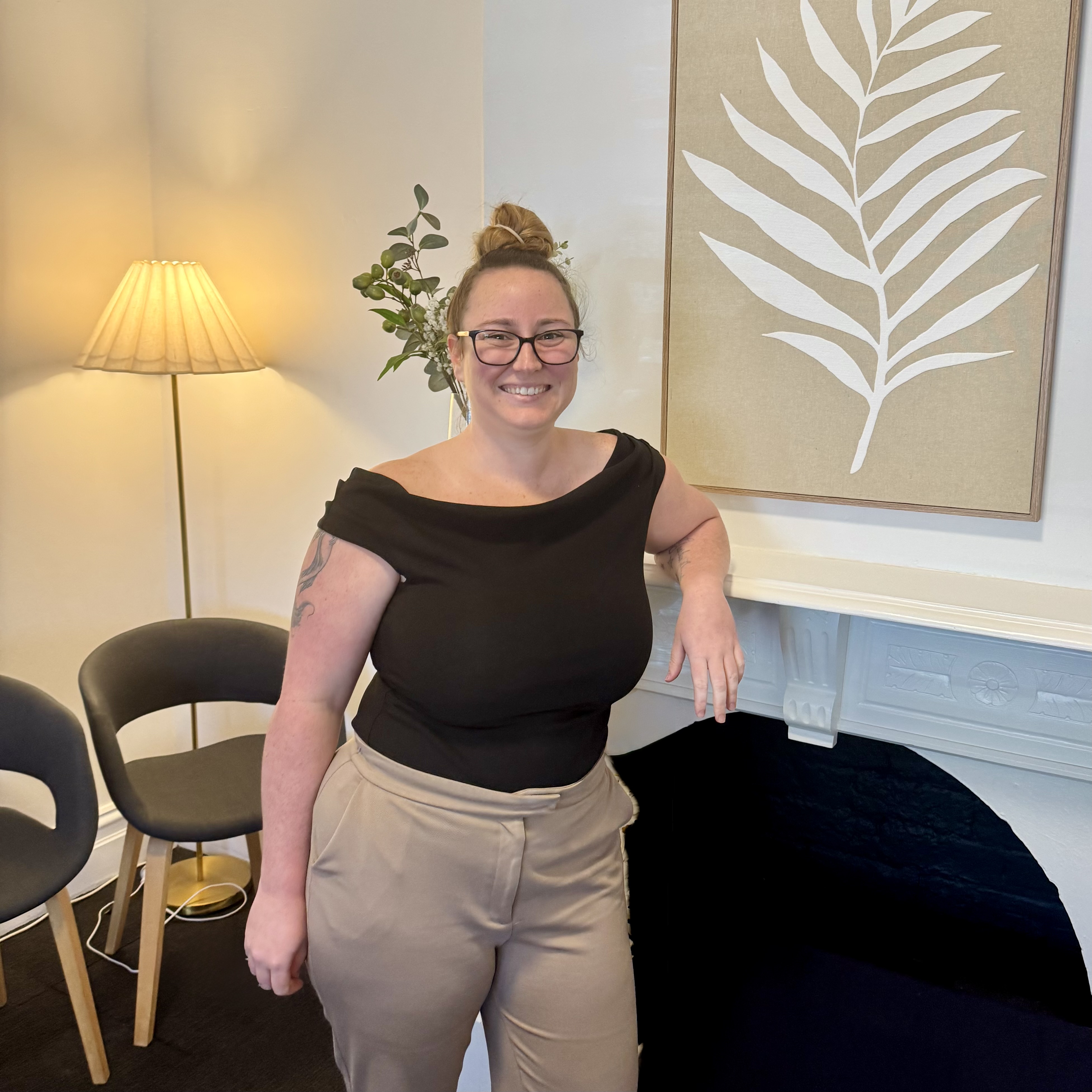 Smiling clinic manager with glasses in a black top and beige pants standing by a fireplace in a room.
