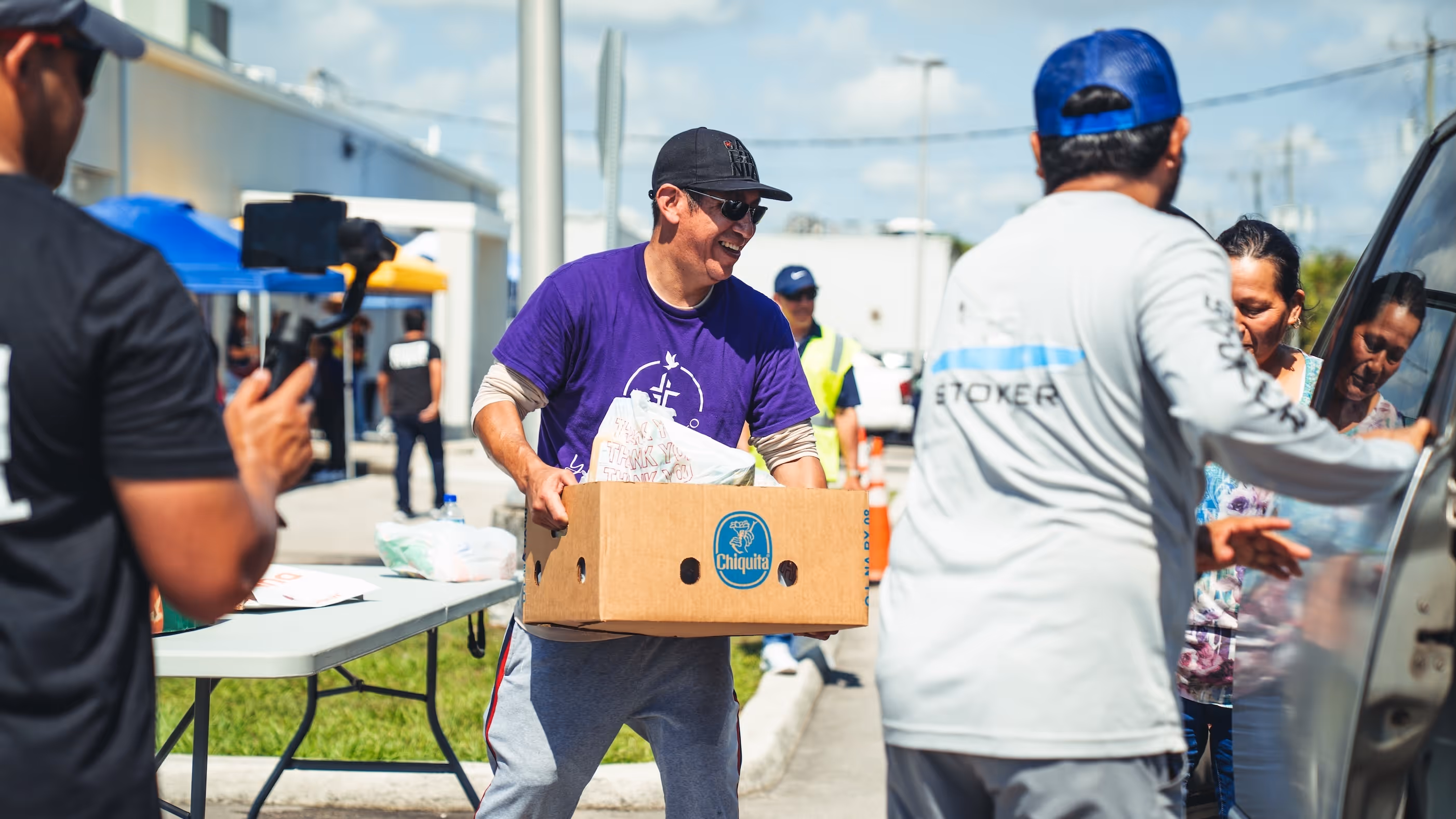 Smiling man in a purple shirt and black cap carrying a Chiquita box filled with items during an outdoor community event.