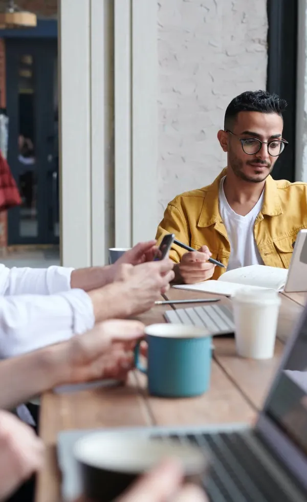 Man in yellow jacket writing in notebook at a table with laptops, coffee mugs, and another person using a smartphone.