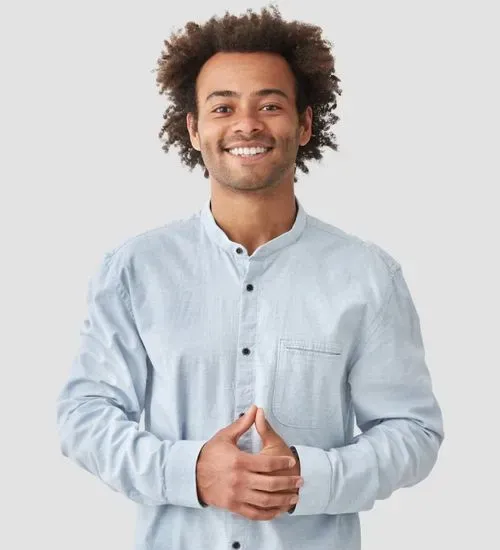 Smiling man with curly hair wearing a light blue button-up shirt, standing with hands clasped.