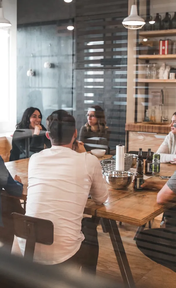 Group of people sitting around a wooden table having a casual meeting with drinks and snacks in a modern office kitchen.