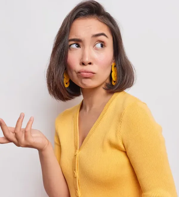 Young woman in yellow sweater and matching earrings looking puzzled with one hand raised in confusion.